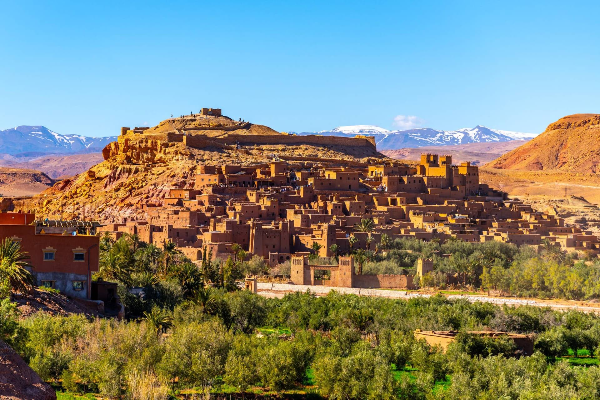 The earthen kasbahs of Aït Benhaddou rising above palm groves in Morocco, with the snow-capped Atlas Mountains behind