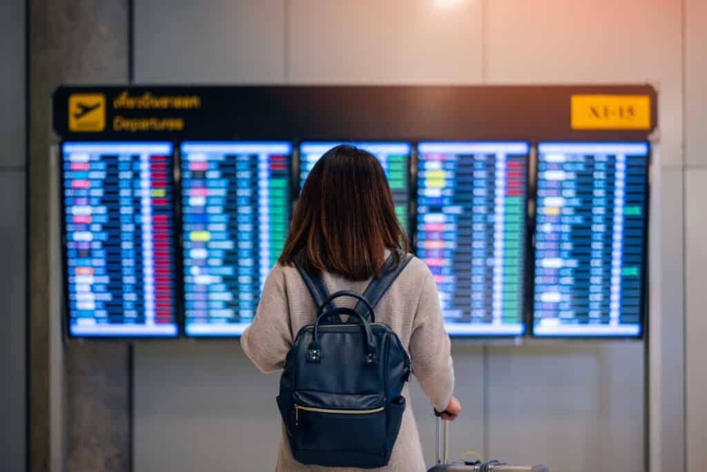 Young woman traveler with backpack and luggage looking at boarding time at departure board at terminal in international airport