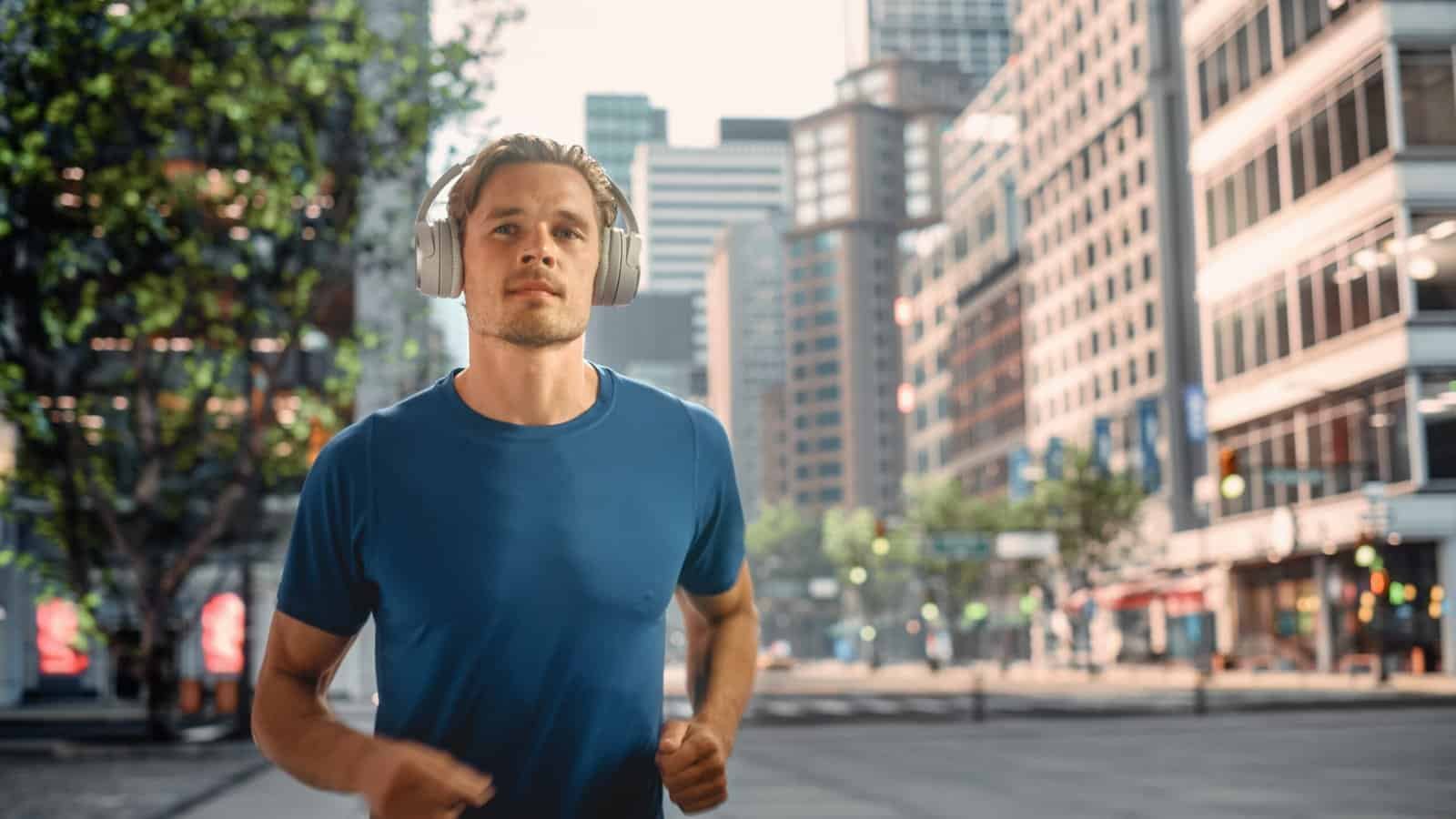 Handsome young man is jogging on a street of a big city center. Male is running in blue t-shirt, wearing wireless headphones with his favourite music playing.
